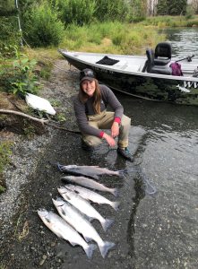 Person crouching in front of a boat on a shore and 6 fish lined up in front of them.