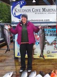 A person smiling outside, while holding a fish in each of their hands, in front of a sign that says Knudson Cove Marina